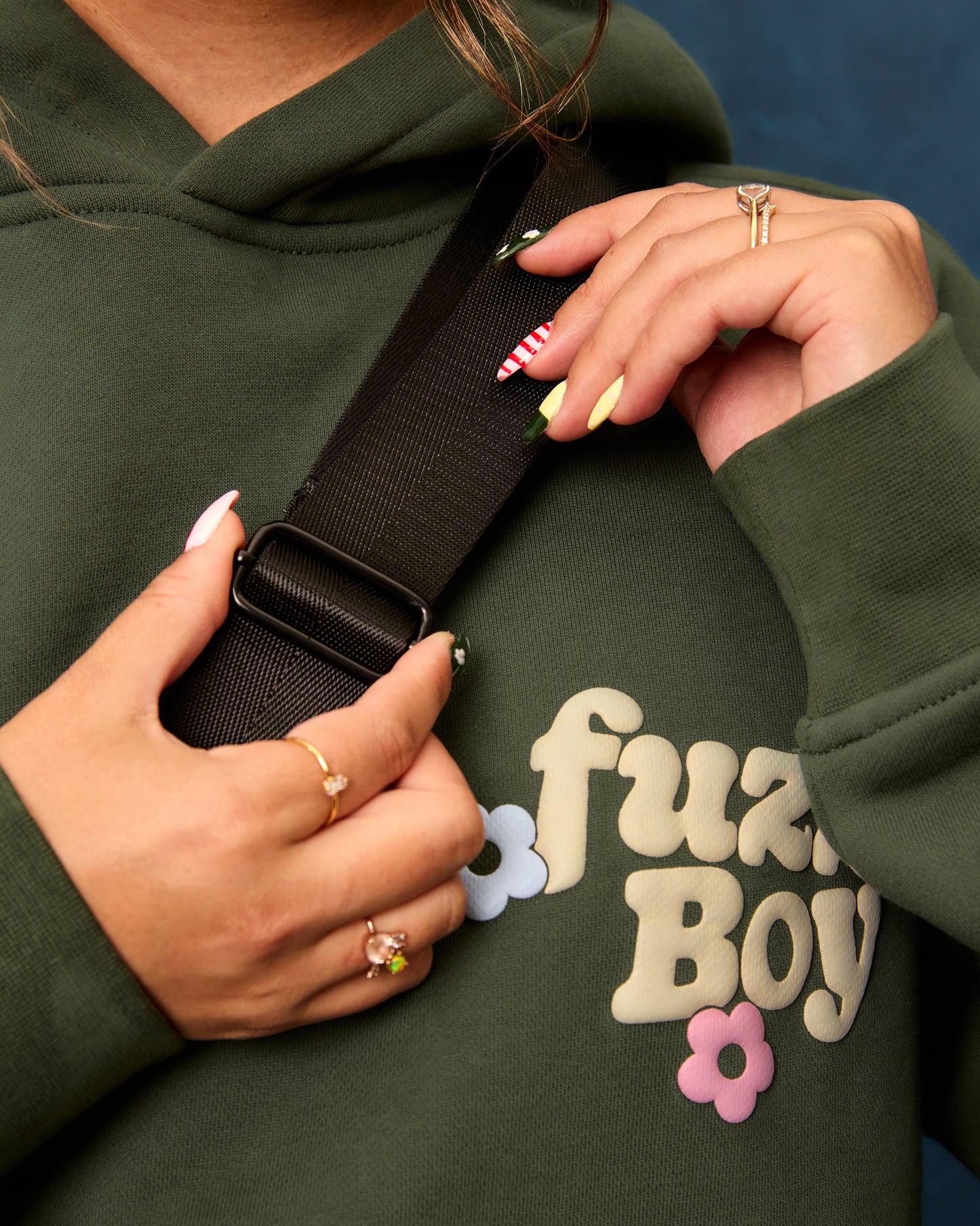 Person wearing a green hoodie with 'Fuzzboy' logo and 3 flowers, demonstrating how to adjust the bag strap.
