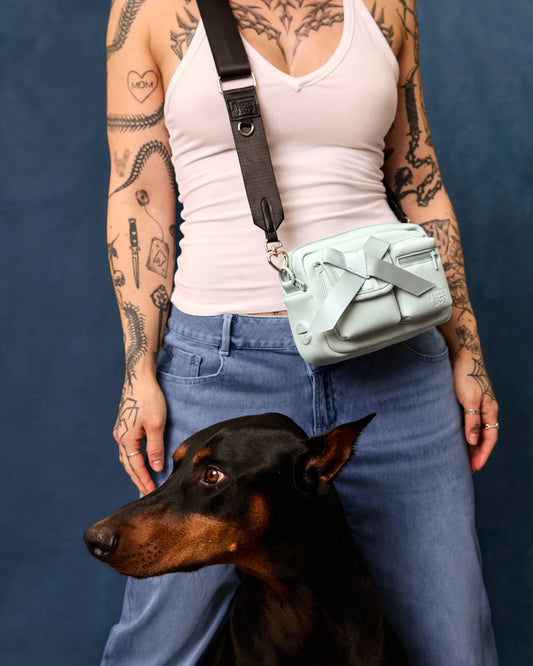 Person wearing a white tank top and blue jeans, and the light blue walking bag worn crossbody. A doberman stands alongside her, in the centre of the frame.