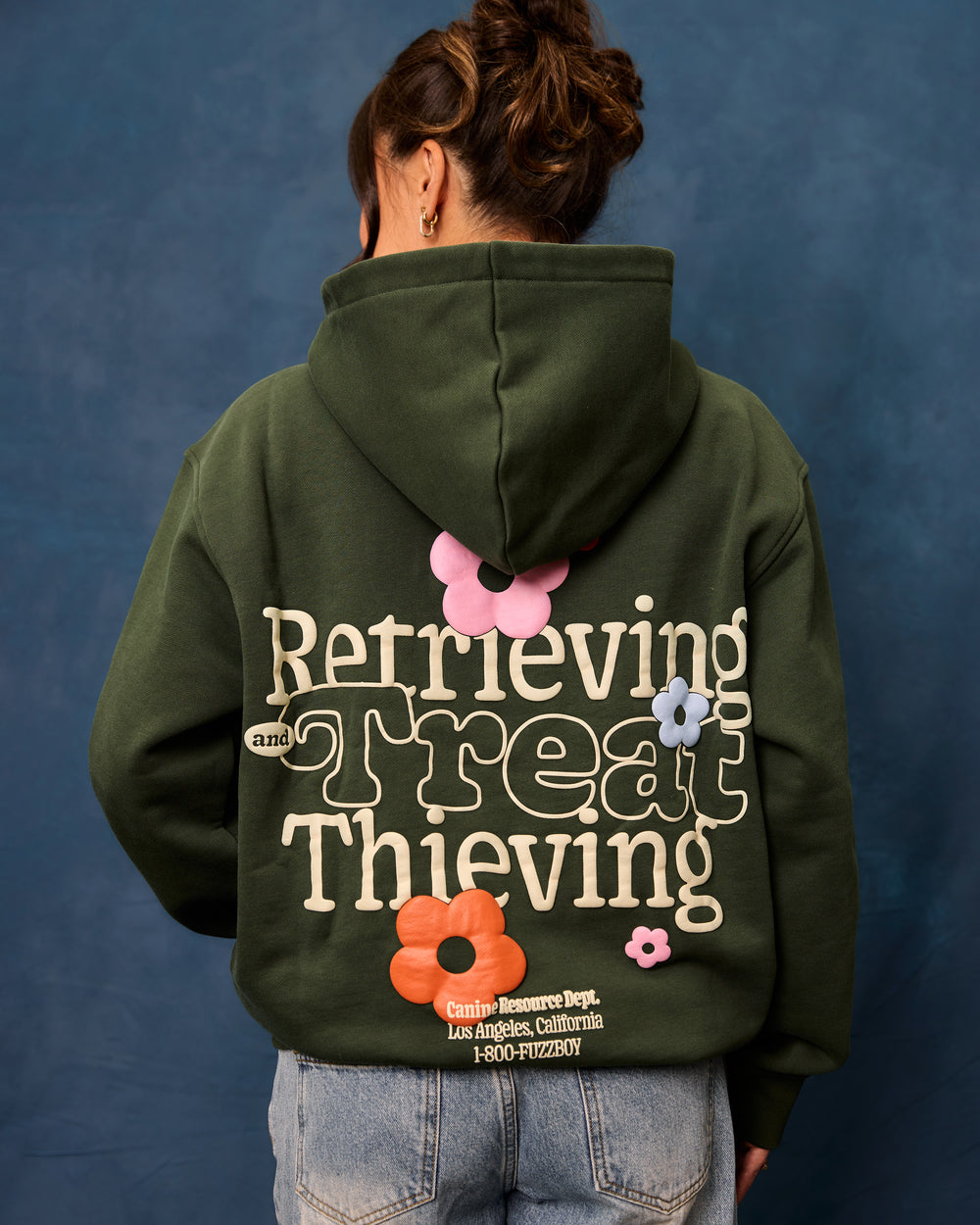 Woman wearing a forest green hoodie with text and floral designs on the back. The text reads "Retrieving and Treat Thieving, Canine Resource Dept. Los Angeles California, 1-800-FUZZBOY.