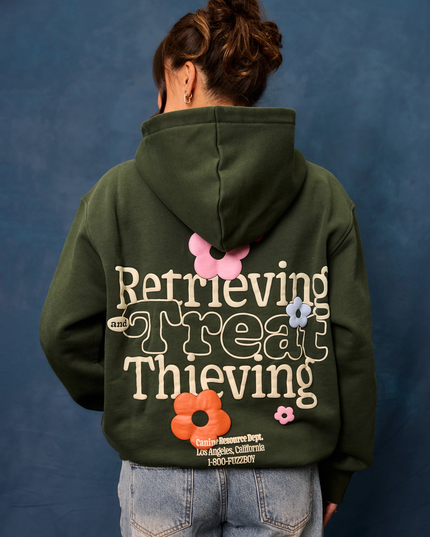 Woman wearing a forest green hoodie with text and floral designs on the back. The text reads "Retrieving and Treat Thieving, Canine Resource Dept. Los Angeles California, 1-800-FUZZBOY.