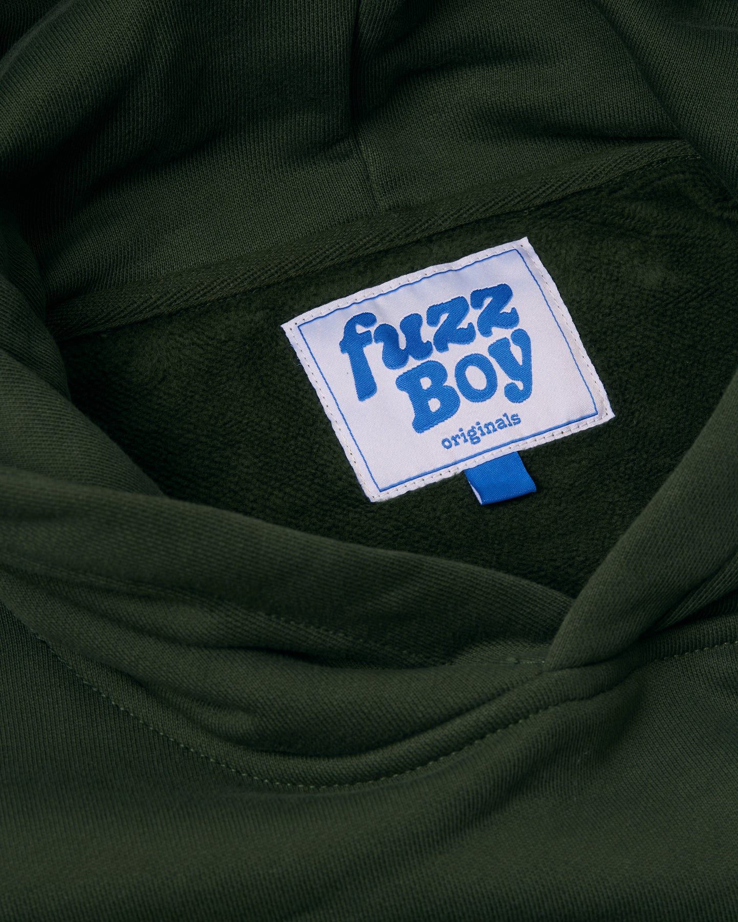 Close-up of the inside tag of the forest green hoodie. White tag reads 'Fuzzboy Originals' in blue text.