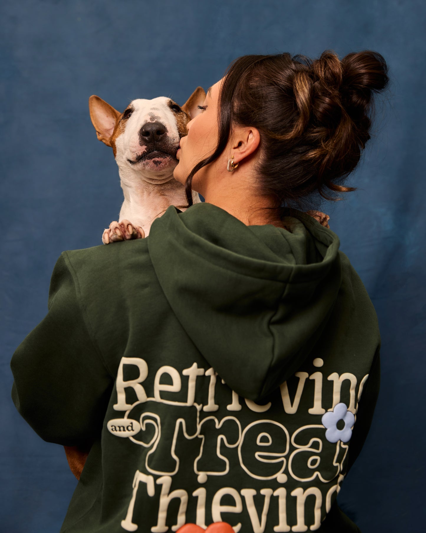 Woman wearing a forest green hoodie with text graphic, holding a mini bull terrier against a blue background.