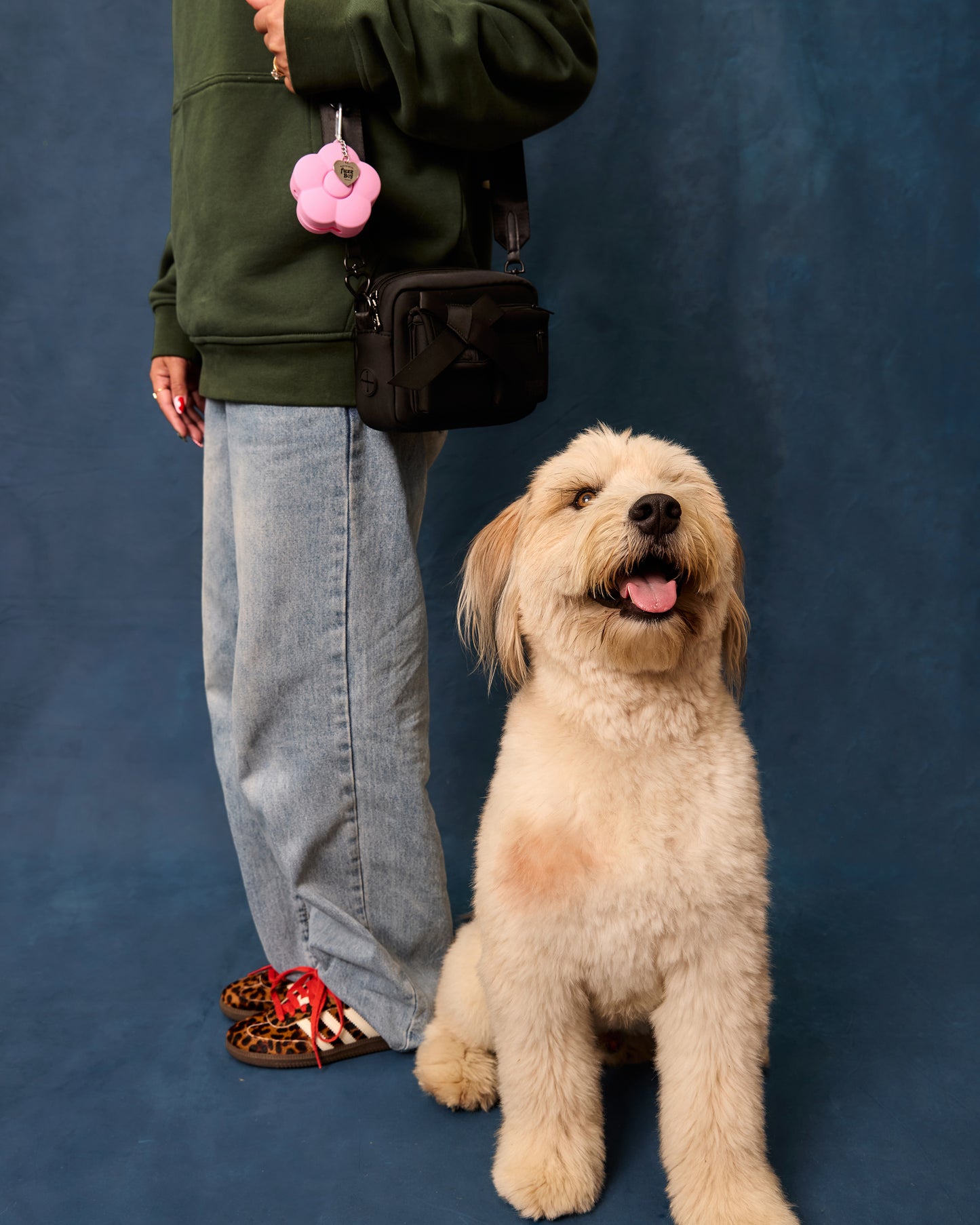 Woman wearing black walking bag on shoulder, with pink flower-shaped treat pouch attached. She poses beside a smiling dog.